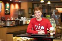 一个大学生坐在咖啡店里，拿着咖啡杯和笔记本电脑，穿着申博太阳城的衬衫，面带微笑